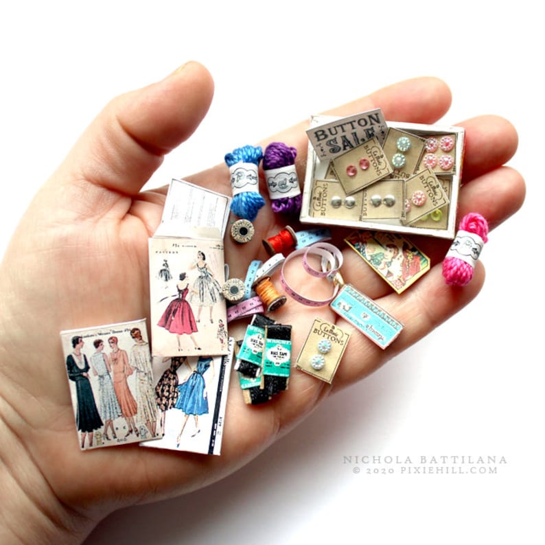 Op de afbeelding: Een verzameling miniatuur naaigerei, waaronder spoelen garen, knopen, een meetlint en vintage naaipatronen. De items zijn gerangschikt op een hand, met een bordje met de tekst "Knopenverkoop".