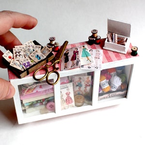 Op de afbeelding: Een miniatuur witte vitrine met roze en witte gestreepte planken gevuld met naai-benodigdheden, waaronder knopen, stofresten, naaipatronen, garen en een schaar. De vitrine heeft een bord met de tekst "4-PLY SOFT KNITTING".