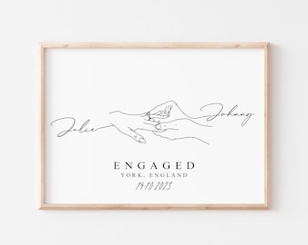 May include: A framed print with a minimalist line drawing of hands, one wearing a ring. The names "Julie" and "Johnny" are written in cursive. Below, the text reads "ENGAGED YORK, ENGLAND 14.10.2023".