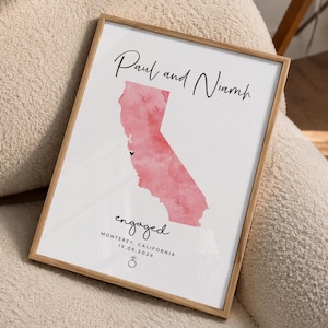 May include: A framed print featuring a watercolour map of California in shades of pink. The text reads Paul and Niamh, engaged, Monterey, California, 19.05.2026, with a small black heart marking a location and a small ring icon at the bottom.