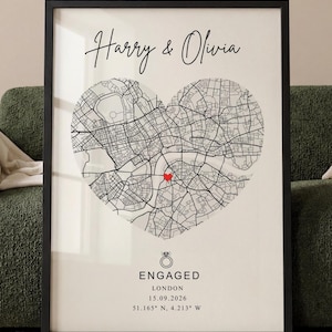 May include: A framed wall art print featuring a black and white street map shaped like a heart. The text reads Harry & Olivia, ENGAGED, LONDON, 15.09.2026, and 51.165° N, 4.213° W. A small red heart marks a location on the map, and a ring icon is included.
