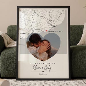 Puede incluir: Un póster vertical enmarcado que muestra un mapa en blanco y negro de Samphire Hoe con un icono de corazón rojo. Debajo del mapa hay una foto en forma de corazón de una pareja, seguida del texto OUR ENGAGEMENT, los nombres Oliver & Gaby y la fecha 26.09.2026.