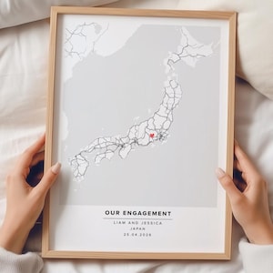May include: A framed map print of Japan in a light wood frame. The map features a light gray background with black outlines of the country and a small red heart icon. Text reads: OUR ENGAGEMENT, LIAM AND JESSICA, JAPAN, 25.04.2026, in black font.