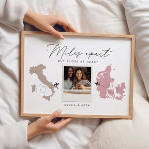 Peut inclure: Un cadre en bois clair contenant une photo de deux femmes, avec des contours de cartes de l'Italie et du Danemark. Le texte dit "Miles apart but close at heart" et les noms "Olivia & Asta".