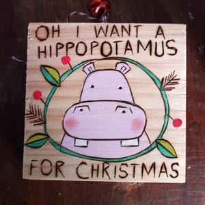 May include: A wooden Christmas ornament with the text "Oh I want a hippopotamus for Christmas" and a cartoon illustration of a pink hippopotamus with green leaves and red berries.