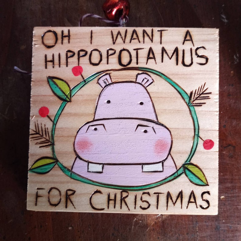 May include: A wooden Christmas ornament with the text "Oh I want a hippopotamus for Christmas" and a cartoon illustration of a pink hippopotamus with green leaves and red berries.