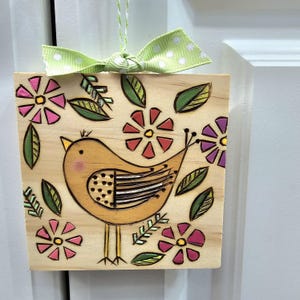 May include: Wooden square with a hand-painted bird design. The bird is yellow with black detailing, surrounded by pink, red, and purple flowers and green leaves. A green and white polka dot ribbon is tied at the top. A decorative wall hanging.