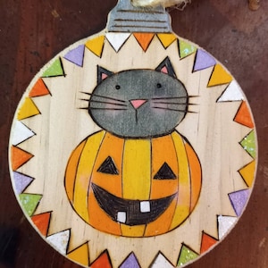 May include: A wooden Halloween ornament with a black cat peeking out from inside a carved pumpkin. The ornament is decorated with a colorful border of triangles.
