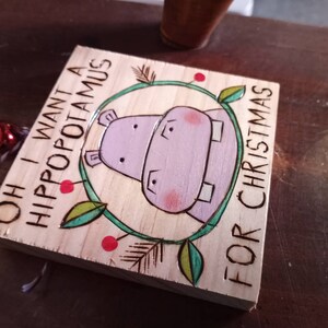 May include: A wooden sign with a hand-painted illustration of a purple hippopotamus with red cheeks. The sign reads "Oh I want a hippopotamus for Christmas".