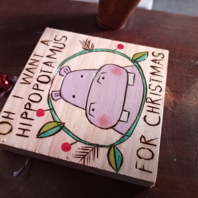 May include: A wooden sign with a hand-painted illustration of a purple hippopotamus with red cheeks. The sign reads "Oh I want a hippopotamus for Christmas".