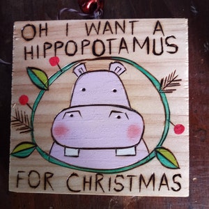 May include: A wooden Christmas ornament with a hand-painted illustration of a pink hippopotamus with the text "OH I WANT A HIPPOPOTAMUS FOR CHRISTMAS".