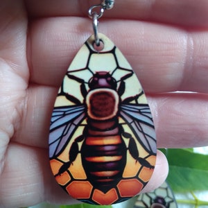 May include: A pair of teardrop-shaped earrings with a bee and honeycomb design. The bee is brown, yellow, and black with purple wings. The honeycomb is orange and yellow.