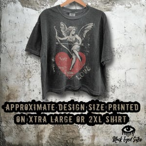 May include: Dark gray t-shirt featuring a distressed graphic of Cupid with a bow and arrow, standing on a red heart with the word "LOVE". The shirt has short sleeves and a crew neck. The text on the shirt reads "APPROXIMATE DESIGN SIZE PRINTED ON XTRA LARGE OR 2XL SHIRT".