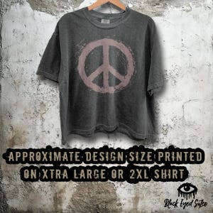 May include: A dark gray t-shirt with a faded pink peace sign graphic. The shirt is hanging on a wooden hanger against a textured wall. The text at the bottom reads "APPROXIMATE DESIGN SIZE PRINTED ON XTRA LARGE OR 2XL SHIRT."
