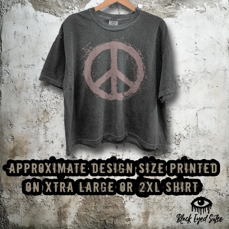 May include: A dark gray t-shirt with a faded pink peace sign graphic. The shirt is hanging on a wooden hanger against a textured wall. The text at the bottom reads "APPROXIMATE DESIGN SIZE PRINTED ON XTRA LARGE OR 2XL SHIRT."
