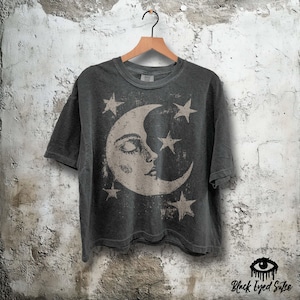 May include: Dark gray t-shirt featuring a crescent moon with a face and several stars. The graphic is in a faded, vintage style. The shirt is hanging on a wooden hanger against a textured wall.