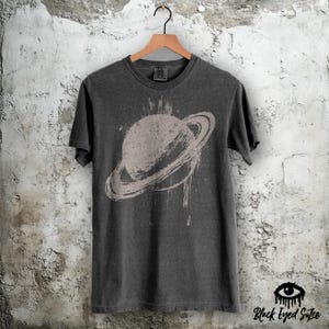 May include: Dark gray t-shirt featuring a distressed graphic of the planet Saturn with its rings. The design is in a lighter gray color, creating a vintage look. The shirt is hanging on a wooden hanger.