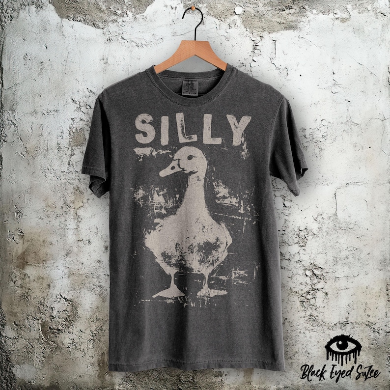 Op de afbeelding: Donkergrijs t-shirt met het woord "SILLY" boven een verweerde afbeelding van een eend. Het shirt hangt aan een houten hanger tegen een getextureerde muur. De merknaam "Black Eyed Sutee" staat rechtsonder.