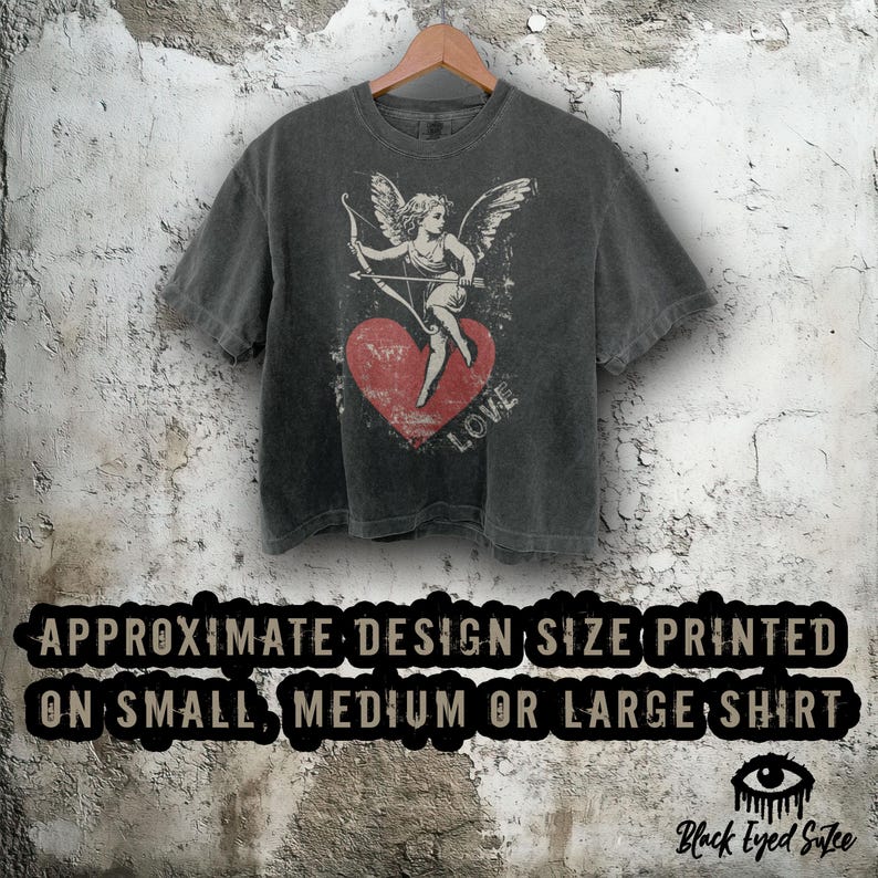 May include: Dark gray t-shirt with a distressed print of Cupid aiming an arrow at a red heart. The word "LOVE" is printed on the heart. The shirt is hanging on a wooden hanger against a textured wall. The text on the bottom says "APPROXIMATE DESIGN SIZE PRINTED ON SMALL, MEDIUM OR LARGE SHIRT".