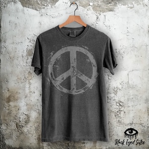 May include: Dark gray t-shirt with a large, distressed peace symbol in the center. The shirt is hanging on a wooden hanger against a textured wall. The brand name "Black Eyed Sutee" is printed in black at the bottom right.