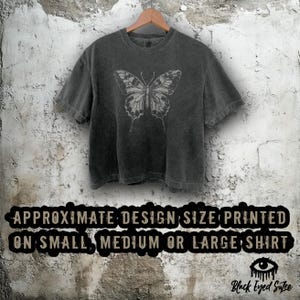 Puede incluir: Camiseta gris oscuro con un dise&ntilde;o de mariposa gris claro. La mariposa es detallada. La camiseta est&aacute; colgada en una percha de madera contra una pared texturizada. El texto en la camiseta dice "APPROXIMATE DESIGN SIZE PRINTED ON SMALL, MEDIUM OR LARGE SHIRT."