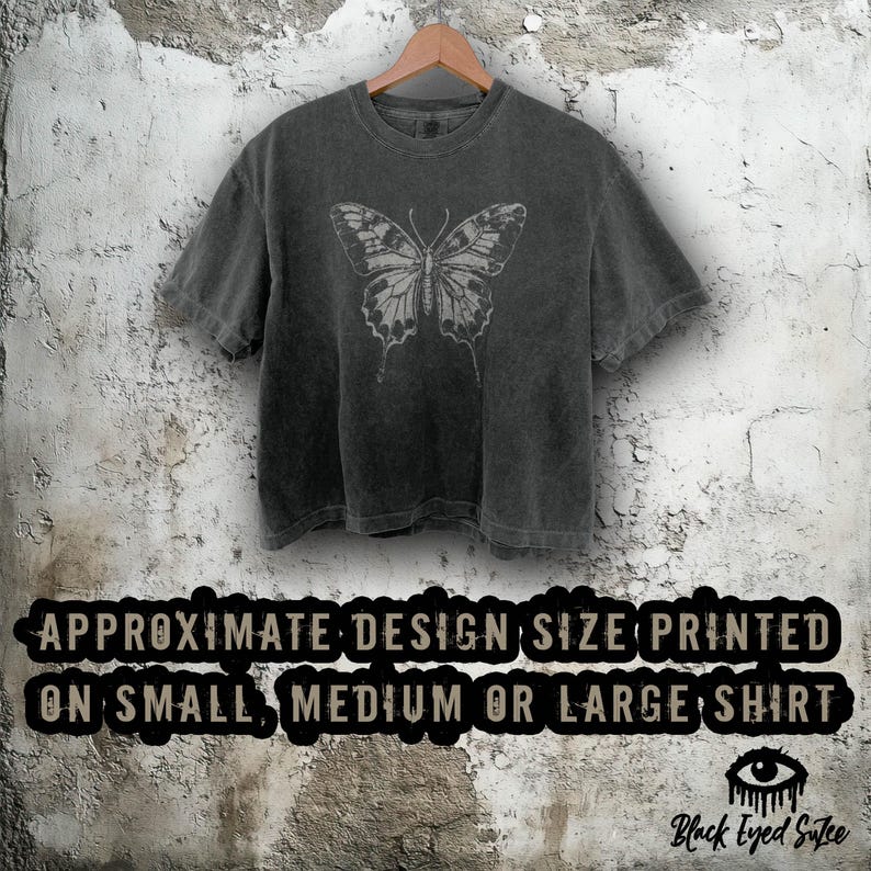 Puede incluir: Camiseta gris oscuro con un dise&ntilde;o de mariposa gris claro. La mariposa es detallada. La camiseta est&aacute; colgada en una percha de madera contra una pared texturizada. El texto en la camiseta dice "APPROXIMATE DESIGN SIZE PRINTED ON SMALL, MEDIUM OR LARGE SHIRT."