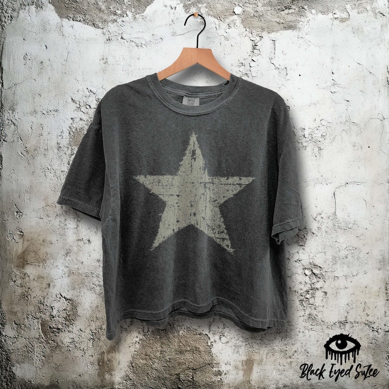 May include: Dark grey t-shirt with a large, distressed white star graphic. The shirt is hanging on a wooden hanger against a textured, off-white wall. The shirt has short sleeves and a crew neck.