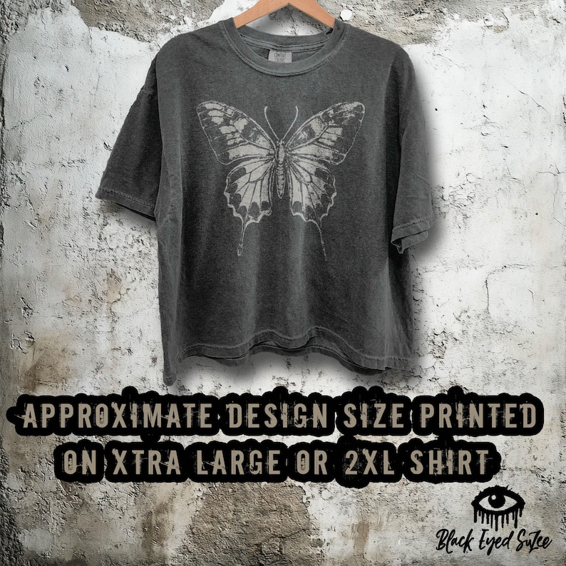 Puede incluir: Camiseta gris oscuro con un gran dise&ntilde;o de mariposa detallado en blanco. La camiseta est&aacute; colgada en una percha de madera contra una pared texturizada blanquecina. El texto en la parte inferior de la imagen dice: "APPROXIMATE DESIGN SIZE PRINTED ON XTRA LARGE OR 2XL SHIRT."