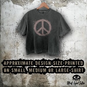 May include: Dark gray t-shirt with a faded pink peace sign graphic. The shirt is hanging on a wooden hanger against a textured wall. Text at the bottom reads "APPROXIMATE DESIGN SIZE PRINTED ON SMALL, MEDIUM OR LARGE SHIRT."