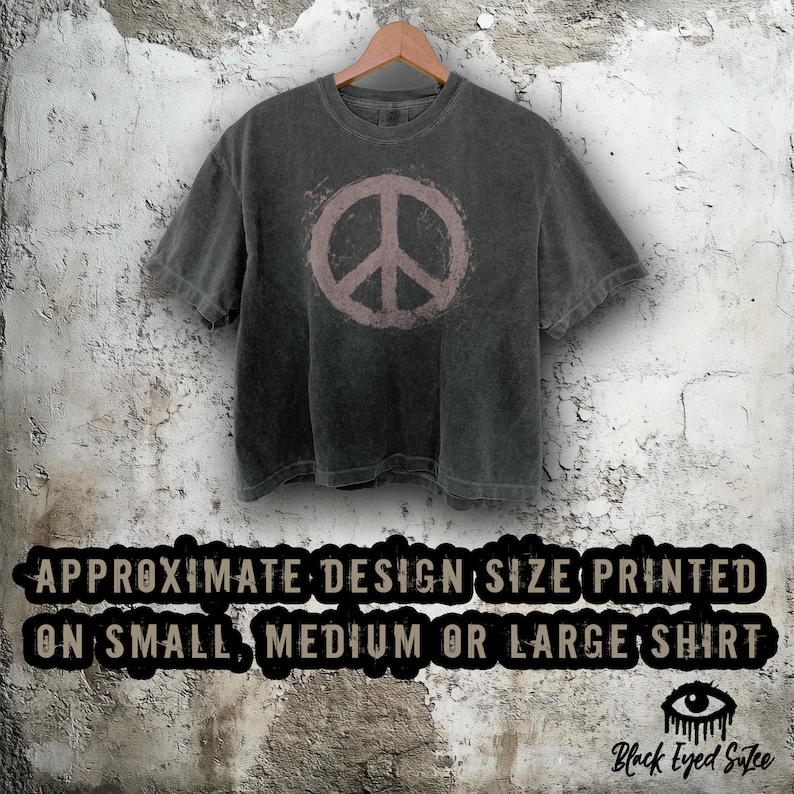 May include: Dark gray t-shirt with a faded pink peace sign graphic. The shirt is hanging on a wooden hanger against a textured wall. Text at the bottom reads "APPROXIMATE DESIGN SIZE PRINTED ON SMALL, MEDIUM OR LARGE SHIRT."