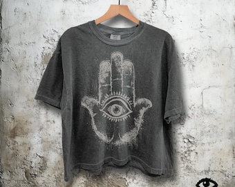 May include: Dark grey t-shirt with a distressed white Hamsa hand graphic featuring an eye in the center. The shirt is hanging on a wooden hanger against a textured wall. The brand logo "Black Eyed Sufee" is visible at the bottom right.