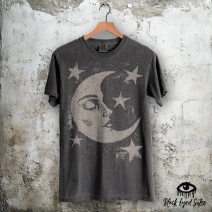 May include: A charcoal grey short-sleeved t-shirt featuring a large, distressed-style graphic of a crescent moon with a human-like face, surrounded by four five-pointed stars. The shirt is displayed on a wooden hanger against a textured wall.