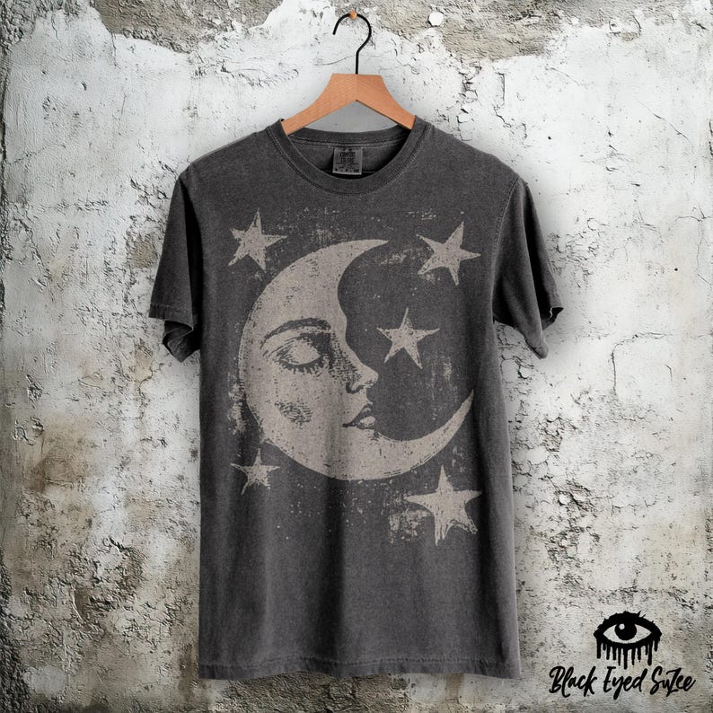 May include: A charcoal grey short-sleeved t-shirt featuring a large, distressed-style graphic of a crescent moon with a human-like face, surrounded by four five-pointed stars. The shirt is displayed on a wooden hanger against a textured wall.