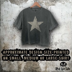 May include: A charcoal gray t-shirt with a distressed white star graphic. The shirt is hanging on a wooden hanger against a textured, off-white wall. The text at the bottom reads "APPROXIMATE DESIGN SIZE PRINTED ON SMALL, MEDIUM OR LARGE SHIRT".