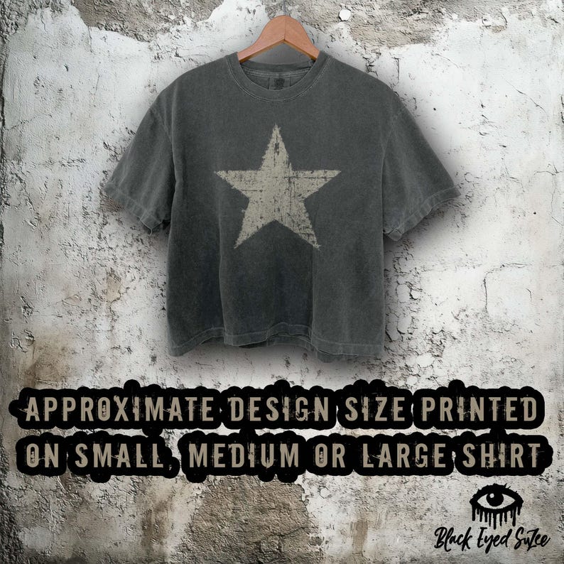 May include: A charcoal gray t-shirt with a distressed white star graphic. The shirt is hanging on a wooden hanger against a textured, off-white wall. The text at the bottom reads "APPROXIMATE DESIGN SIZE PRINTED ON SMALL, MEDIUM OR LARGE SHIRT".