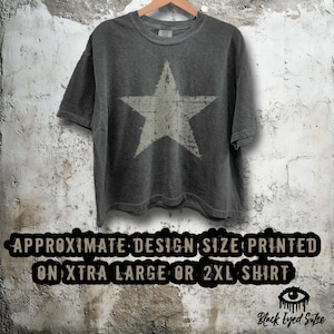 May include: Dark gray t-shirt with a large, distressed white star graphic. The shirt is hanging on a wooden hanger against a textured wall. Text at the bottom reads "APPROXIMATE DESIGN SIZE PRINTED ON XTRA LARGE OR 2XL SHIRT."
