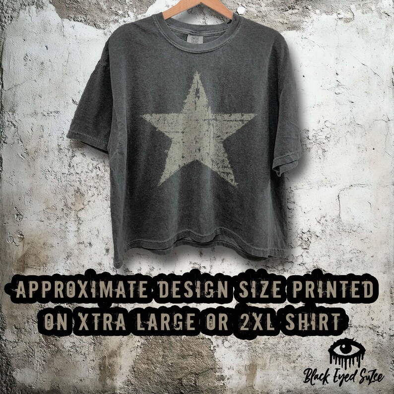May include: Dark gray t-shirt with a large, distressed white star graphic. The shirt is hanging on a wooden hanger against a textured wall. Text at the bottom reads "APPROXIMATE DESIGN SIZE PRINTED ON XTRA LARGE OR 2XL SHIRT."