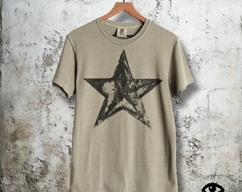 May include: A light olive green t-shirt with a large, distressed black star graphic. The shirt is hanging on a wooden hanger against a textured, off-white wall. The brand name "Black Eyed Sylee" is visible in the bottom right corner.
