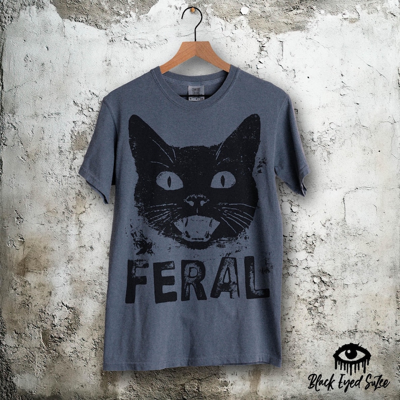 May include: A gray t-shirt featuring a large black cat face with the word "FERAL" printed below. The cat has wide eyes and an open mouth. The shirt is hanging on a wooden hanger against a textured wall.
