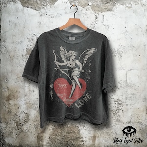 May include: Dark gray t-shirt featuring a distressed graphic of Cupid with a bow and arrow, standing on a red heart. The word "LOVE" is printed below the heart. The shirt is hanging on a wooden hanger.