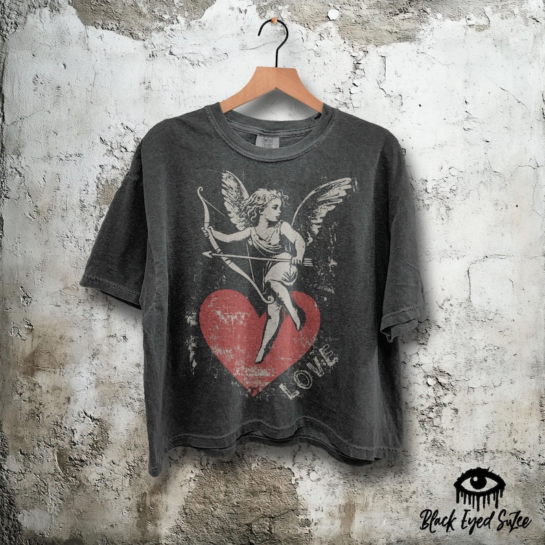 May include: Dark gray t-shirt featuring a distressed graphic of Cupid with a bow and arrow, standing on a red heart. The word "LOVE" is printed below the heart. The shirt is hanging on a wooden hanger.