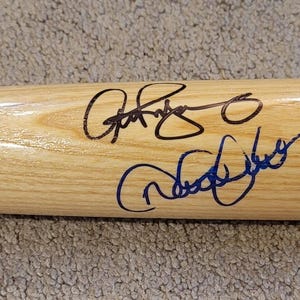 Puede incluir: Un bate de béisbol de madera clara con múltiples firmas en tinta negra. El bate muestra la veta natural de la madera. Las firmas varían en estilo y tamaño. El bate está colocado horizontalmente.