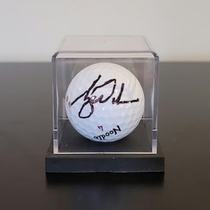 May include: A white golf ball with a black signature, presented in a clear acrylic case on a black base. The golf ball features the word "Noodle" and the number "7". The case is displayed on a dark surface.