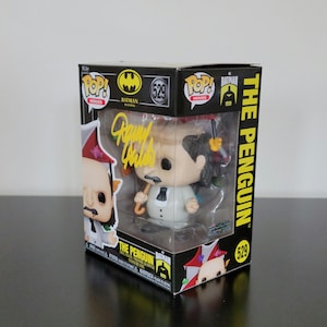 Może przedstawiać: Figurka winylowa Funko Pop! Pingwina z Batman Returns, w oryginalnym pudełku. Figurka ma na sobie biały garnitur i trzyma parasol. Pudełko jest czarne z żółtymi akcentami i zawiera logo Batmana oraz numer 529.