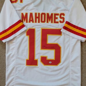 Puede incluir: Camiseta de fútbol blanca con rayas rojas y amarillas en las mangas. La parte posterior de la camiseta tiene el nombre "MAHOMES" y el número "15" en rojo con un contorno amarillo. La camiseta está firmada con tinta negra.