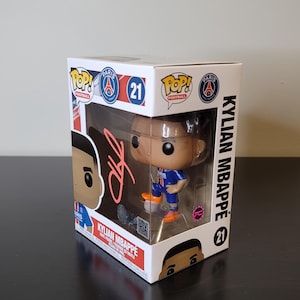May include: A white box with a clear plastic window showing a Funko Pop! figure of Kylian Mbappé wearing a blue and white football shirt with the number 7 on the back. The box has the words "Pop! Football" and "Paris" on it. The figure is holding a football. The box also has the number 21 on it.