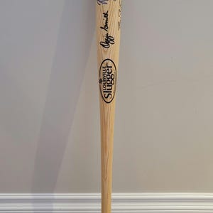 May include: A wooden Louisville Slugger baseball bat with the text "Genuine Ozzie Smith" and his signature in blue ink. The bat is light brown and has a rounded end.