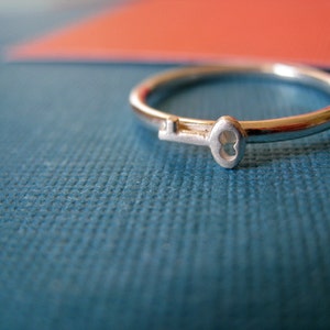 May include: A silver ring with a keyhole design. The ring is on a blue textured surface.