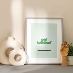 May include: A framed print with the text "get hooked" in green letters on a light green background. The print is sitting on a table with two white ceramic vases and a wooden hand holding a small potted plant.