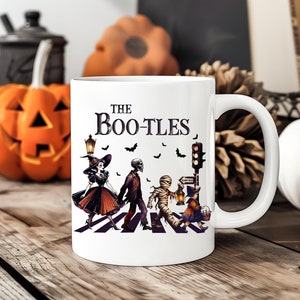 May include: White ceramic mug with the text "The Bootles" and a Halloween-themed illustration. The illustration depicts costumed figures crossing a crosswalk, including a witch, skeleton, and mummy. Black bats and a traffic light are also featured.